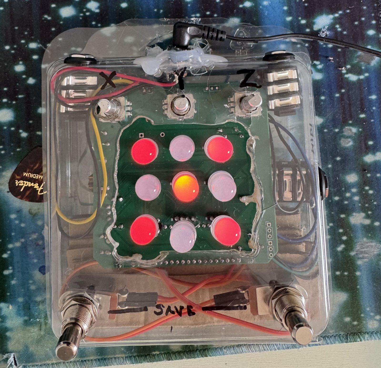The stone's board inside a clear plastic box, x, y and z written over three knobs, SAVE written between two footswitches, its nine lights red and white