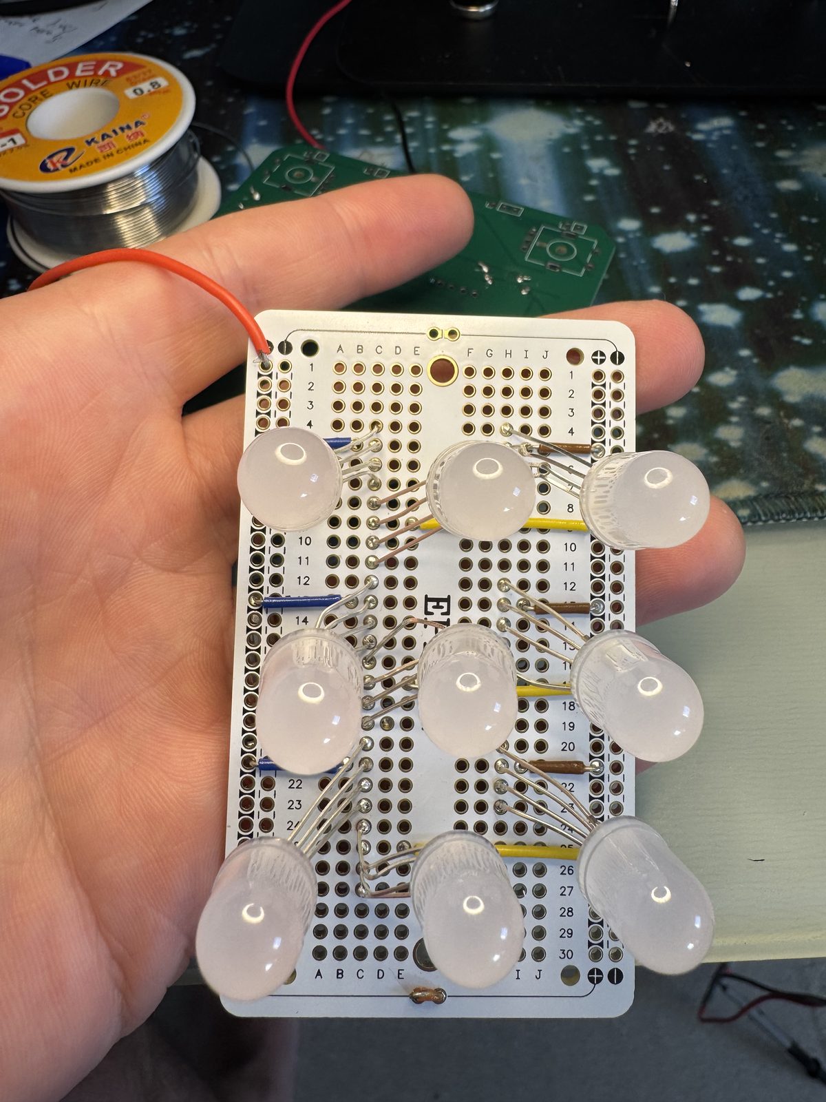 Nine milky round lights in three rows of three, standing in a small perfboard held in a hand, a reel of solder behind