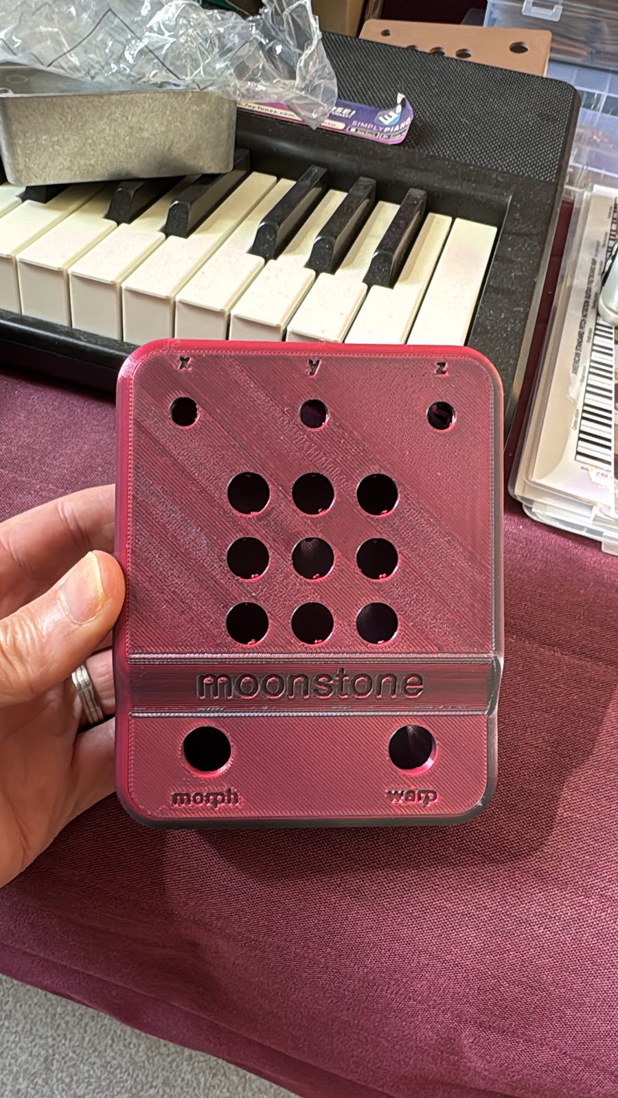 A red printed moonstone face held up in front of a keyboard: x, y and z over three knob holes, nine light holes, the word moonstone, morph and warp