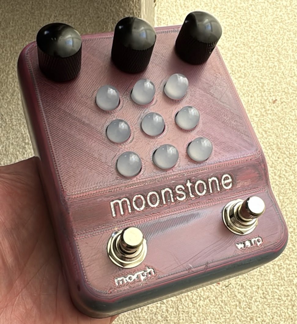 A rose-pink printed moonstone held in a hand: three black knobs, nine milky lights, the word moonstone across its face, and two footswitches marked morph and warp
